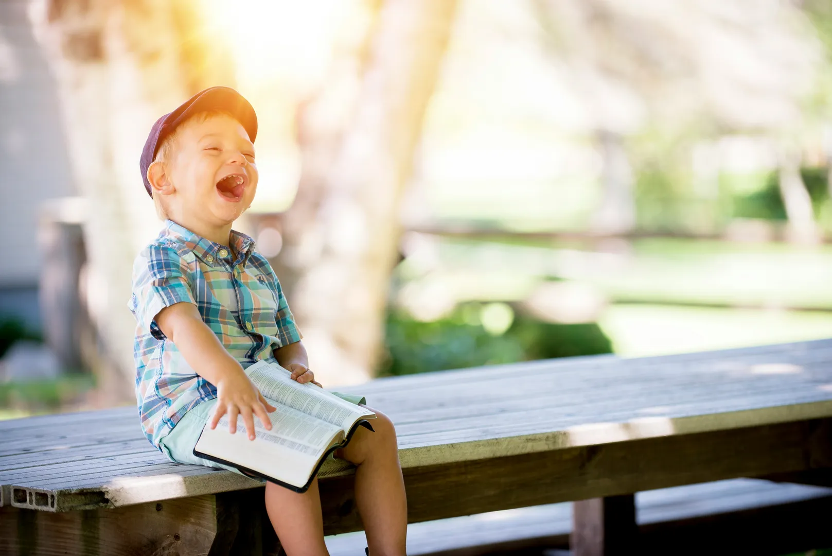 Child laughing, holding open book, sitting on wooden bench.